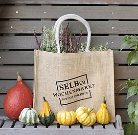 Tasche vor einer Holzwand mit Logo Selber Wochenmarkt, davor Kürbisse und in der Tasche Winterblumen